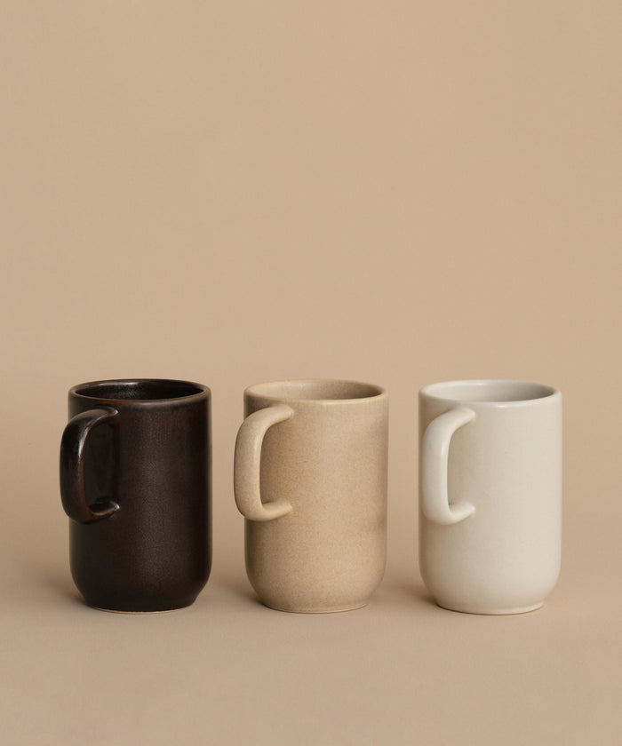 Three Pacific Mugs in black, beige, and white—crafted from stoneware in Portugal—are displayed side by side on a light brown surface with a beige background, showcasing their identical minimalist design.