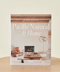The book Pacific Natural At Home stands upright on a wooden surface, its cover featuring a minimalist California living room with neutral tones, wooden beams, and modern furniture.