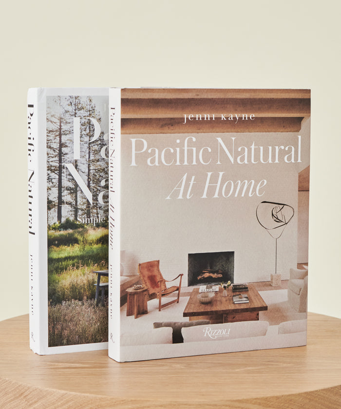 Two books stand upright on a wooden surface. The front book, Pacific Natural At Home, features a neutral-toned California living room cover. A second book is partially visible behind it.