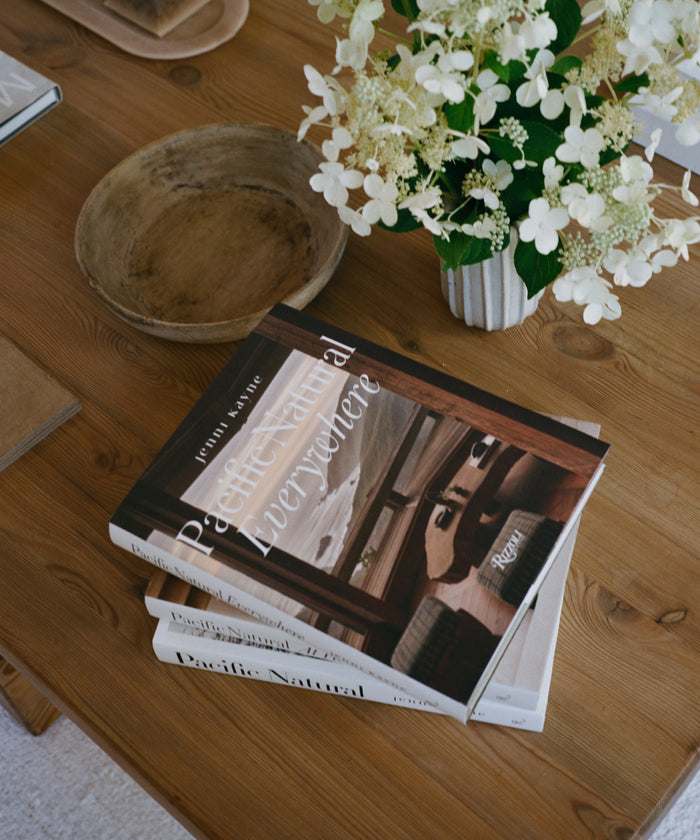 Stack of books on a wooden table with a vase of white flowers and a wooden bowl.