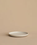 A minimalist, round, white Pacific Salad Plate made of stoneware in Portugal rests empty on a beige surface with a beige background.