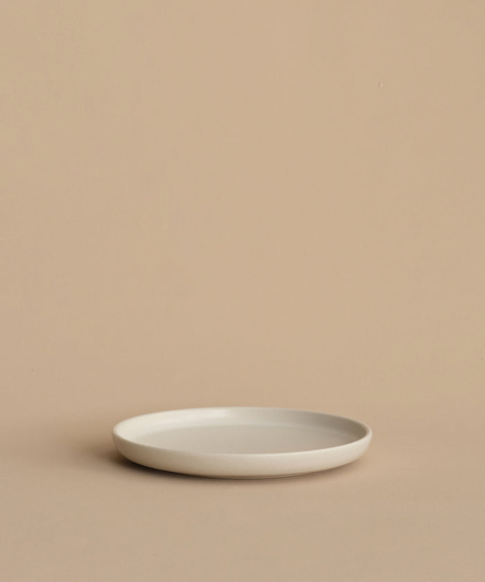 A minimalist, round, white Pacific Salad Plate made of stoneware in Portugal rests empty on a beige surface with a beige background.