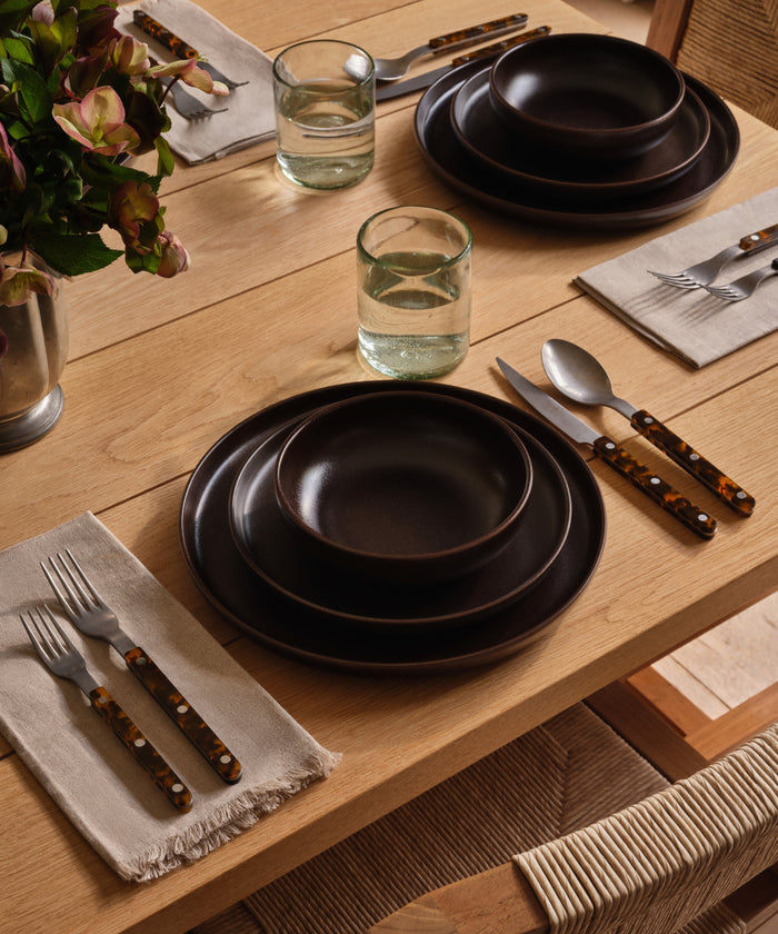 A wooden dining table set with Pacific Salad Plates, brown ceramic bowls, clear water glasses, beige napkins, and brown-handled cutlery. A vase of flowers decorates the tables corner.