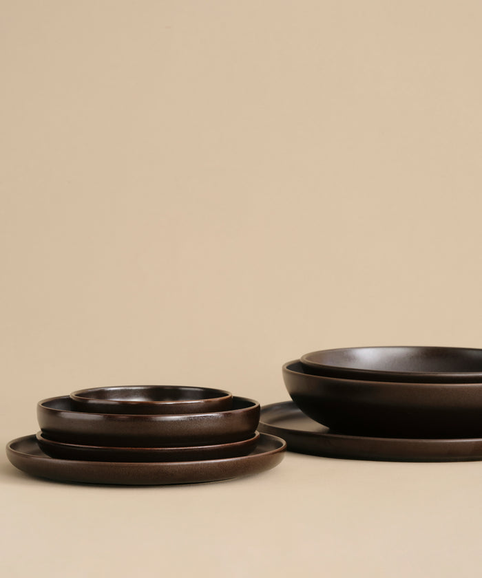 A stack of Pacific Salad Plates in dark brown, arranged on a beige surface with a beige background in two neat piles.