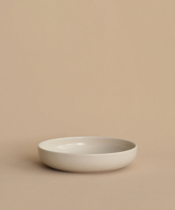 A simple, round, shallow white Pacific Soup Bowl is displayed on a beige surface against a matching background, highlighting its minimalistic, pattern-free elegance.