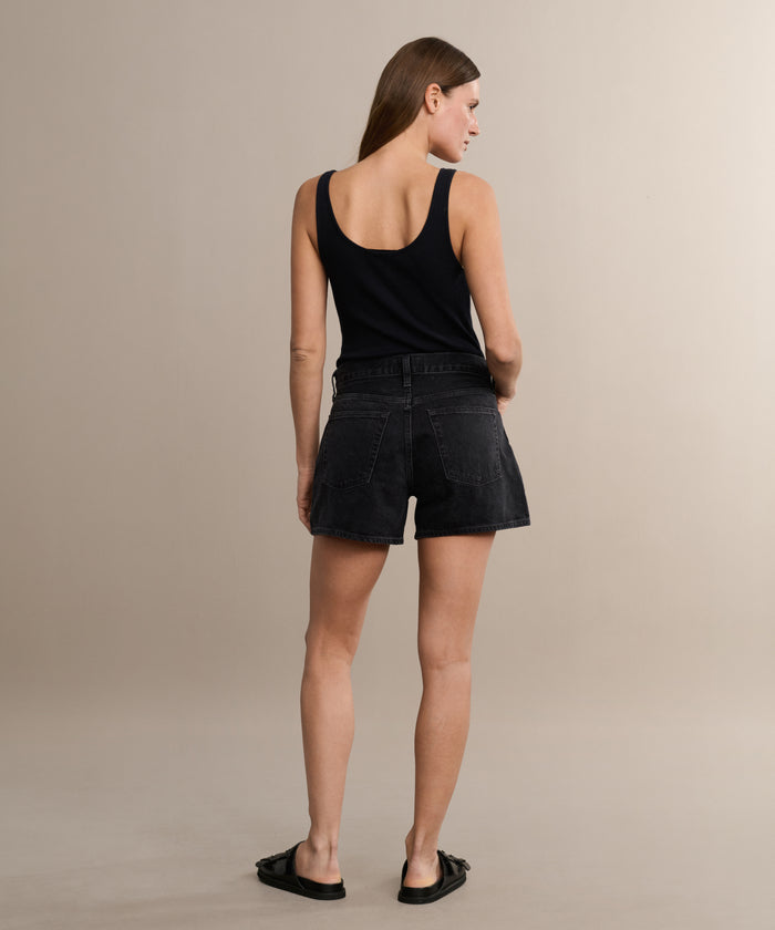 A woman faces away, wearing a black sleeveless top, the Parker Long Short by AGOLDE in a vintage denim fit, and black sandals, set against a plain beige background.