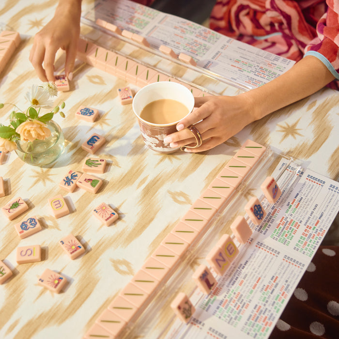 A close-up lifestyle shot of a Mahjong game in progress on a patterned tablecloth, with hands visible holding a cup and arranging tiles. The tiles are light pink with multicolored characters and symbols, and game score sheets are visible in the background.