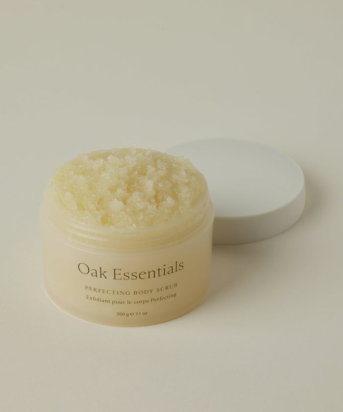 An open jar of Perfecting Body Scrub with its white lid beside it shows the textured, grainy exfoliating formula inside. Set on a neutral background, this hydrating skincare treat is beautifully highlighted.