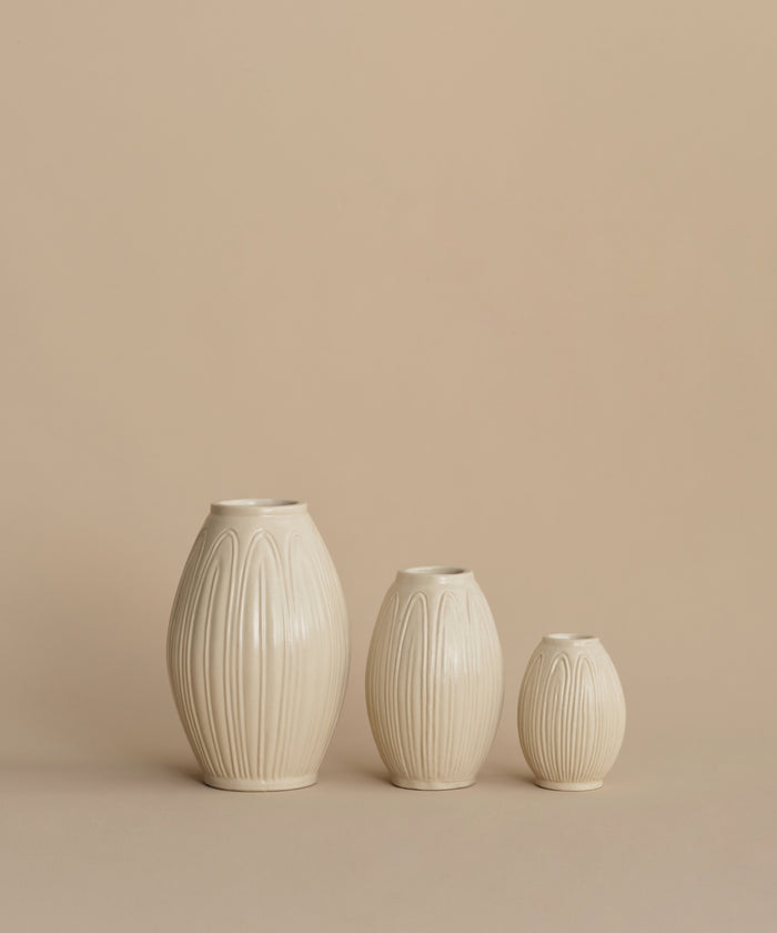 Three ribbed ceramic vases in a warm off-white or cream color, arranged by size against a solid beige background.