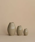 Three ribbed ceramic vases in a light beige-gray glaze, arranged by size against a solid warm beige background.