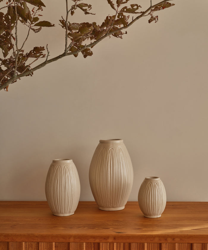 Three ribbed ceramic vases in a warm cream-beige tone, displayed on a wooden surface against a beige wall with dried branches hanging from the top.