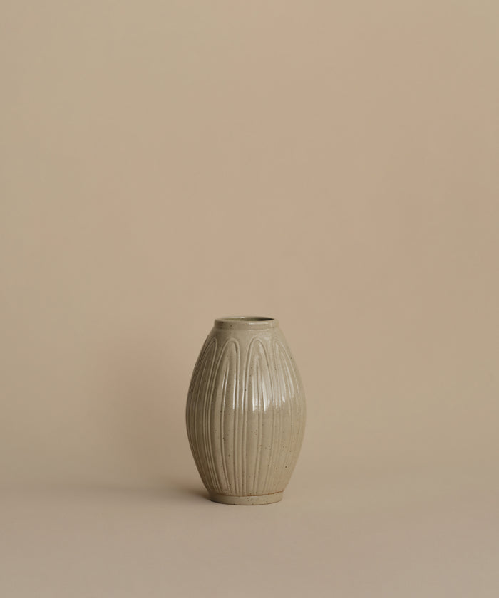 A small ribbed ceramic vase in a light warm gray-beige glaze, centered against a solid beige background.