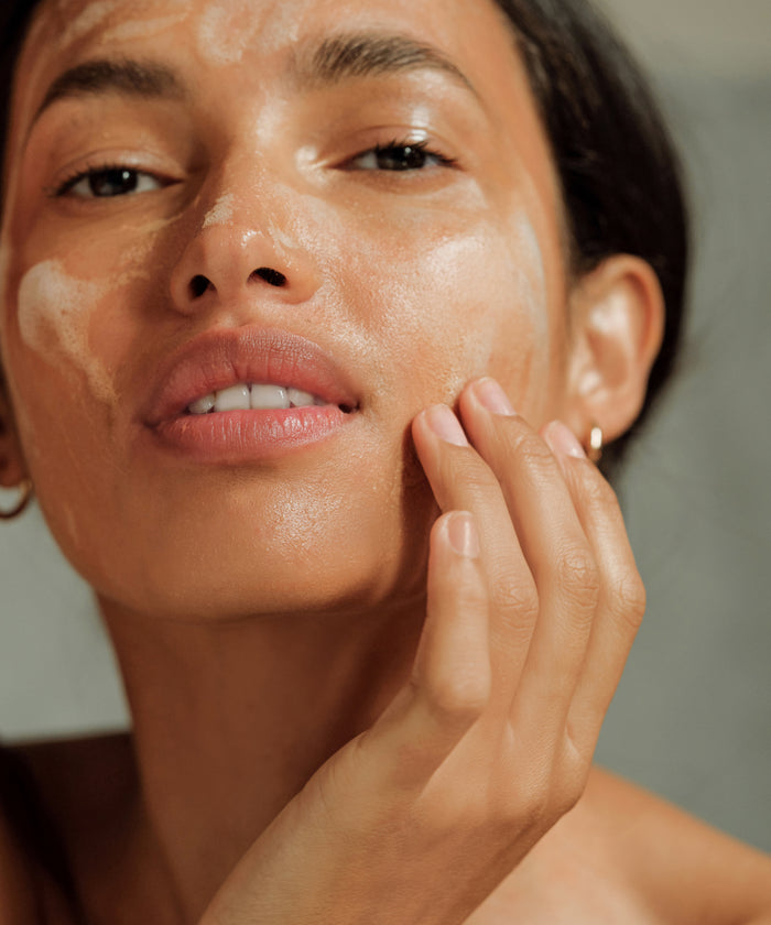 A close-up of a person applying Pure Gel Cleanser to their face, gently touching their cheek. Their dewy, well-lit skin suggests a brighter complexion, while their expression remains neutral.