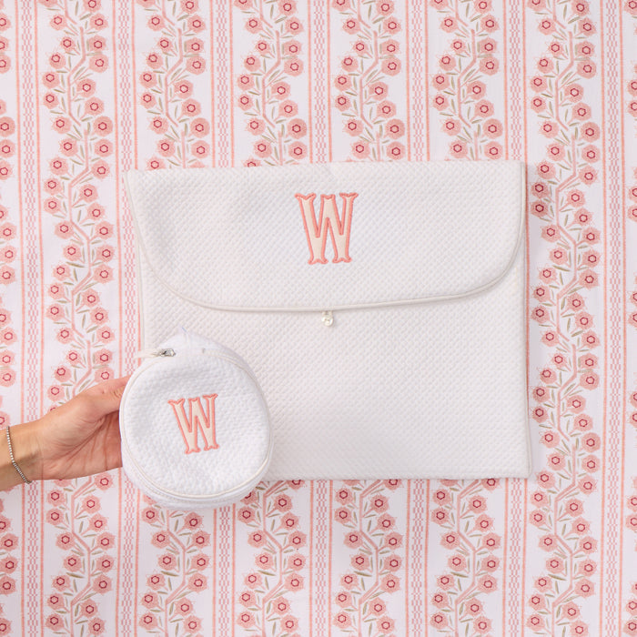 A white textured fabric pouch with a flap closure and a matching round coin pouch held in a hand, both embroidered with a coral-pink monogram letter 'W', set against a pink and white patterned background.