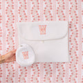 A white textured fabric pouch with a flap closure and a matching round coin pouch held in a hand, both embroidered with a coral-pink monogram letter 'W', set against a pink and white patterned background.