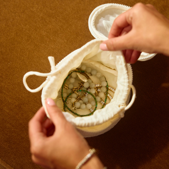 A cream-colored fabric jewelry organizer pouch with drawstrings, shown open with hands holding it to reveal several beaded bracelets inside. The pouch is placed on a textured brown surface.