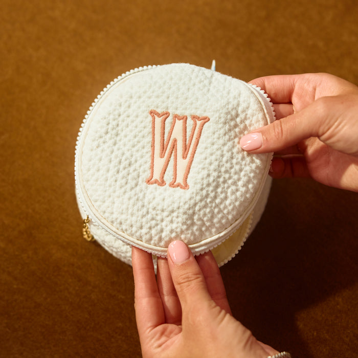A close-up of a round, textured cream-colored zipper pouch with a peach embroidered monogram 'W' on the front, held by two hands against a warm brown background.