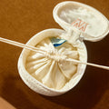 An open round white fabric pouch with a drawstring closure, shown on a warm brown background. A thin wooden stick or tool rests across the opening, and a small blue-and-white packet is visible inside.