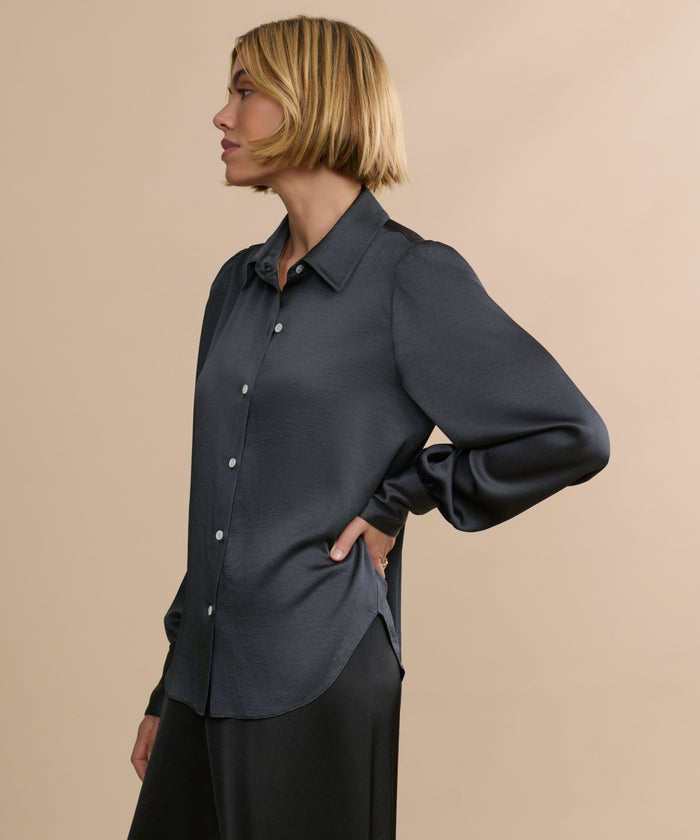 A person with short blonde hair, dressed in a dark gray Remy Shirt featuring subtle puffed sleeves and black pants, stands sideways against a plain beige background.
