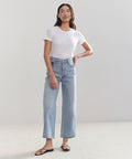 A woman with long dark hair stands confidently in front of a plain background, wearing a white t-shirt, black sandals, and AGOLDE’s Ren Jean in light blue.