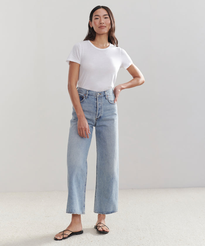 A woman with long dark hair stands confidently in front of a plain background, wearing a white t-shirt, black sandals, and AGOLDE’s Ren Jean in light blue.