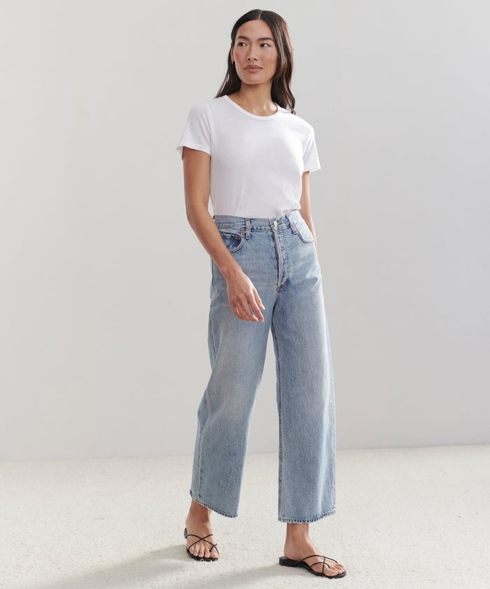A woman with long dark hair stands indoors against a plain background, wearing a white t-shirt, light blue AGOLDE Ren Jean high-waisted jeans, and black sandals, with a relaxed expression.