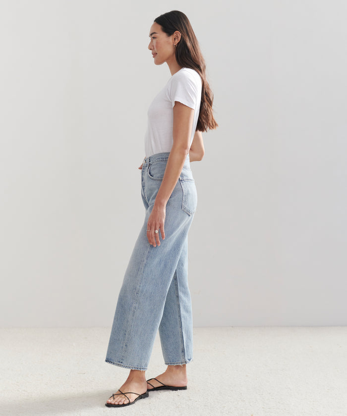 A woman stands in profile against a plain background, wearing a white short-sleeve t-shirt, black sandals, and high-waisted AGOLDE Ren Jean denim jeans in light blue.