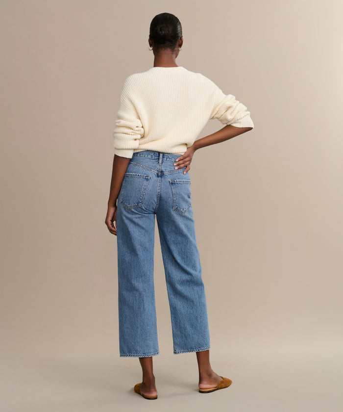 A person faces away, wearing a cream-colored sweater, brown flats, and the Ren jean—high-rise blue jeans with a wide-leg cut—against a plain beige background.