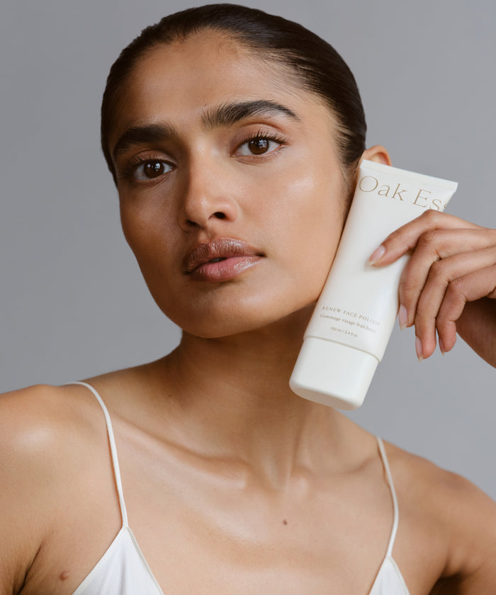A woman with smooth, glowing skin holds a tube of Renew Face Polish near her face, featuring upcycled pineapple exfoliants. She wears a white spaghetti strap top and has a neutral expression against a plain gray background.