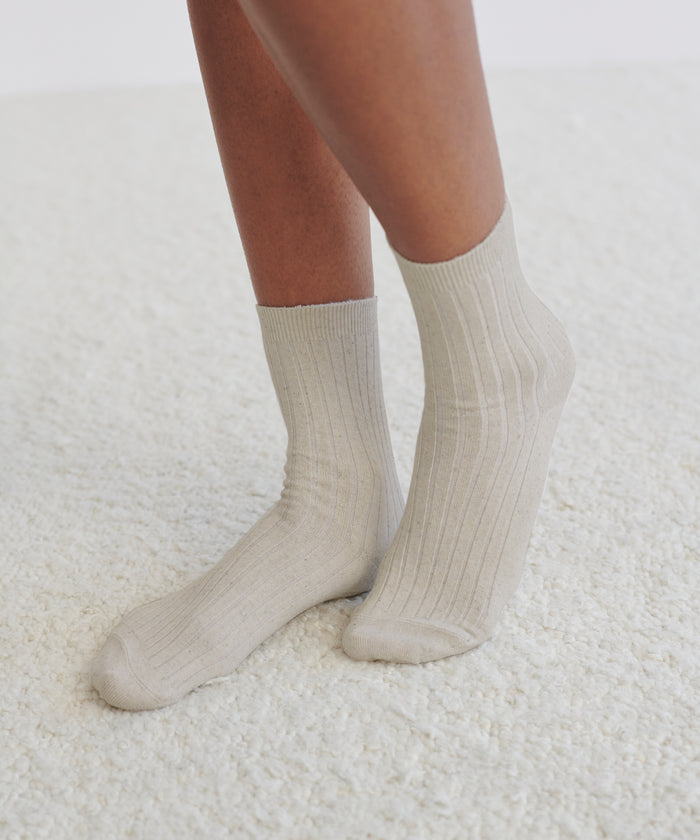 Only the lower legs and feet are visible as a person stands on a white carpeted floor, wearing light beige Rib Socks that reach just above the ankles—a perfect wardrobe staple.