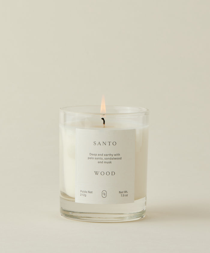 A lit Santo Wood Candle in a clear glass jar with a "SANTO WOOD" label sits against a light cream background. This musky candle blends deep palo santo, sandalwood, and musk. 212g / 7.5oz.