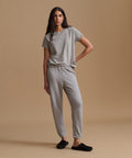 A person wearing light gray heather jogger sweatpants with an elastic waistband and ankle cuffs, paired with a matching gray top and black slip-on slippers, against a warm beige studio background.