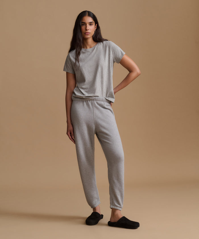 A person wearing light gray heather jogger sweatpants with an elastic waistband and ankle cuffs, paired with a matching gray top and black slip-on slippers, against a warm beige studio background.