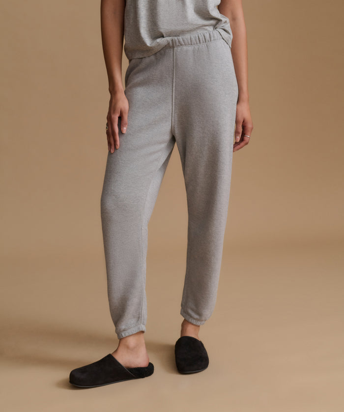 A person wearing light gray heather jogger sweatpants with an elastic waistband and ankle cuffs, paired with a matching gray top and black slip-on slippers, against a warm beige studio background.