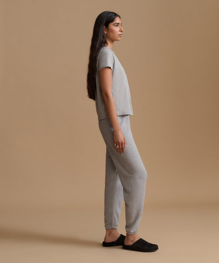 A woman stands in profile wearing a light gray short-sleeve cropped top and matching jogger-style pants, paired with black slip-on shoes, against a plain warm beige studio background.