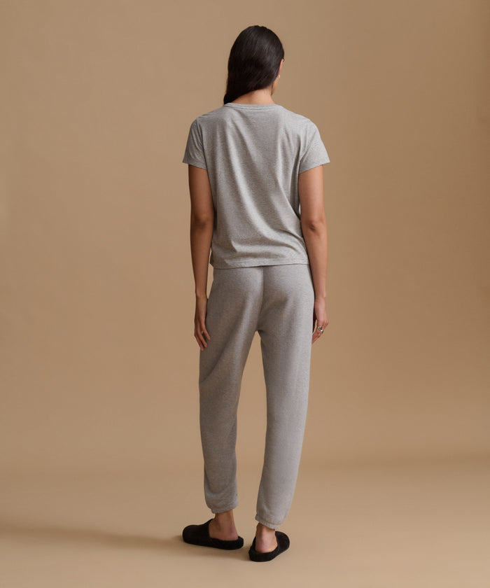 A woman viewed from behind wearing a light gray short-sleeve t-shirt and matching light gray jogger pants, standing against a solid beige studio background.