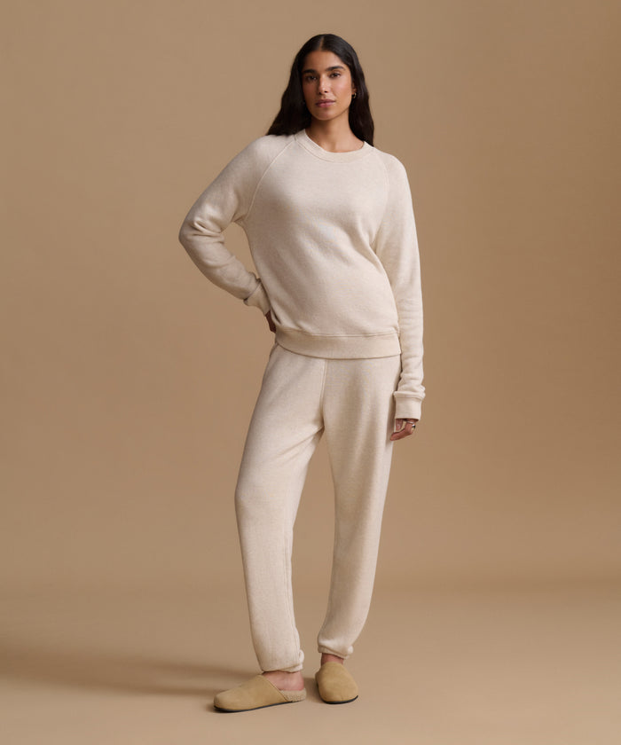 A woman modeling a matching cream knit lounge set consisting of a long-sleeve crewneck sweatshirt and relaxed-fit sweatpants, paired with tan slip-on slippers against a solid beige studio backdrop.
