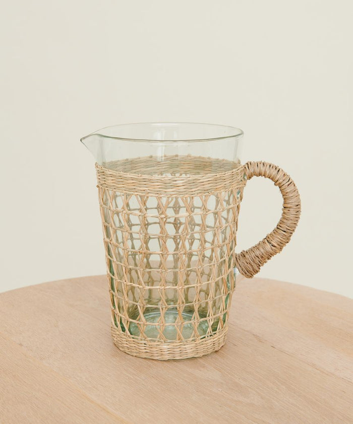 The Seagrass Picnic Pitcher, featuring a woven wicker cover and handle, rests on a light wooden table, bringing natural charm to your tabletop decor against a simple background.
