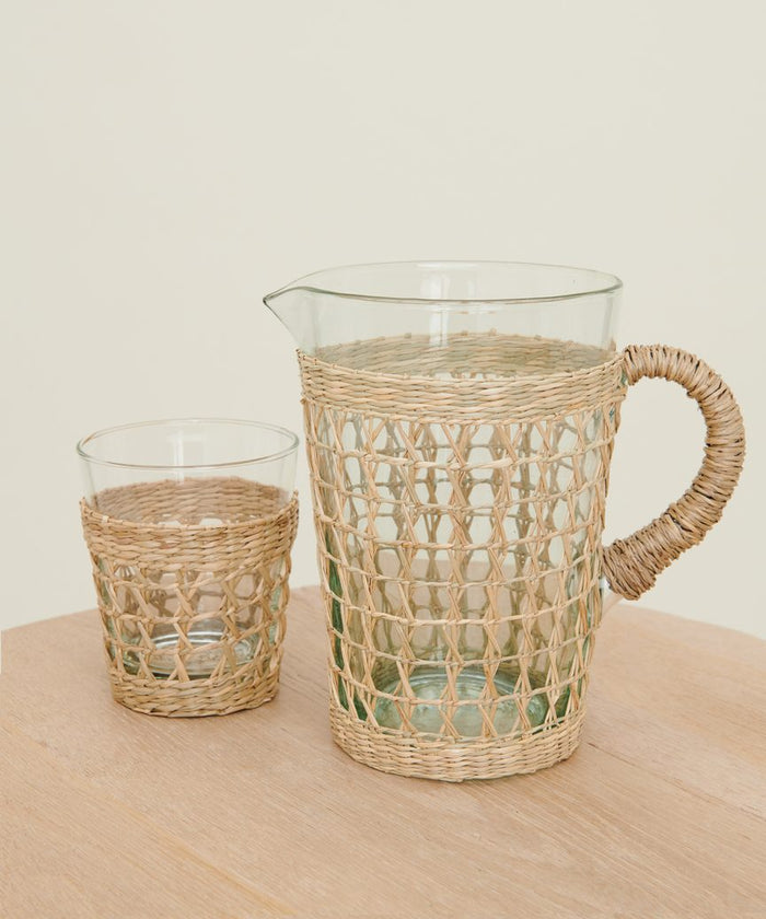 The Seagrass Picnic Pitcher, wrapped in woven wicker, rests on light wood against an off-white background—ideal for an elegant woven tabletop display.