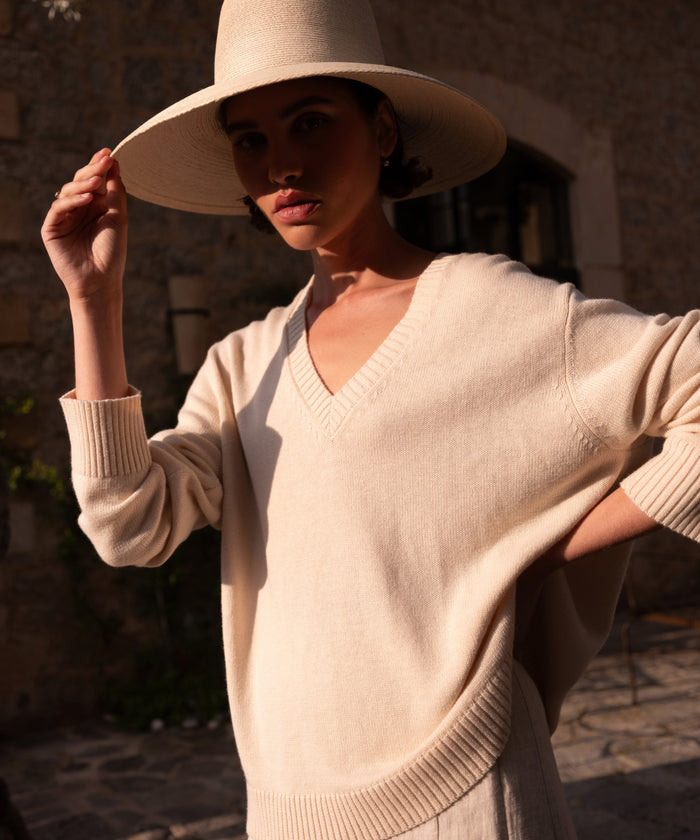 Wearing the Simone V-Neck, a person with a wide-brimmed hat stands outdoors in warm sunlight, their face shaded. Behind them, a stone building completes the scene.