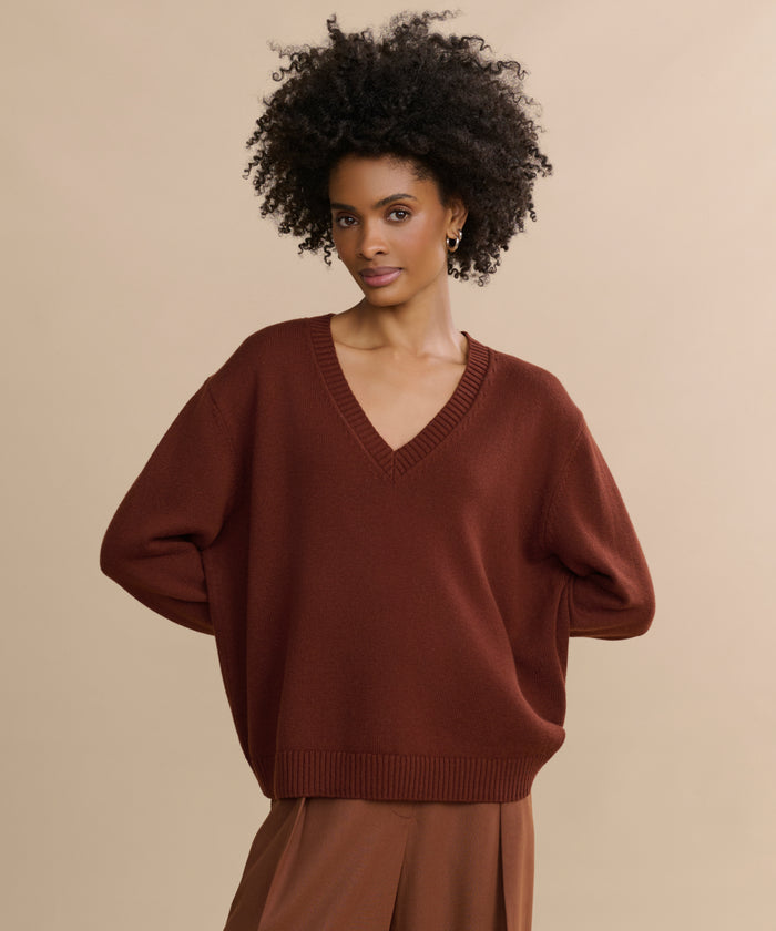 A woman with curly hair wears the Simone V-Neck, a loose-fitting sweater, paired with brown pants. She stands against a plain beige background with her arms behind her back, looking at the camera.