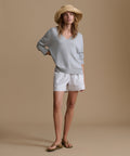 Woman wearing a light blue v-neck sweater with blue and white stripe shorts and a raffia hat against a brown wall.