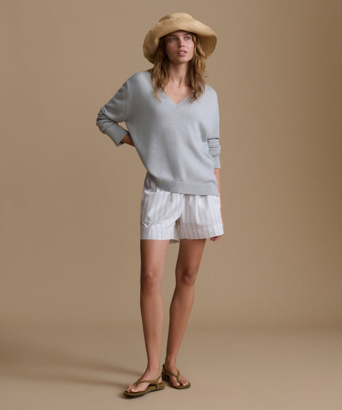 Woman wearing a light blue v-neck sweater with blue and white stripe shorts and a raffia hat against a brown wall.