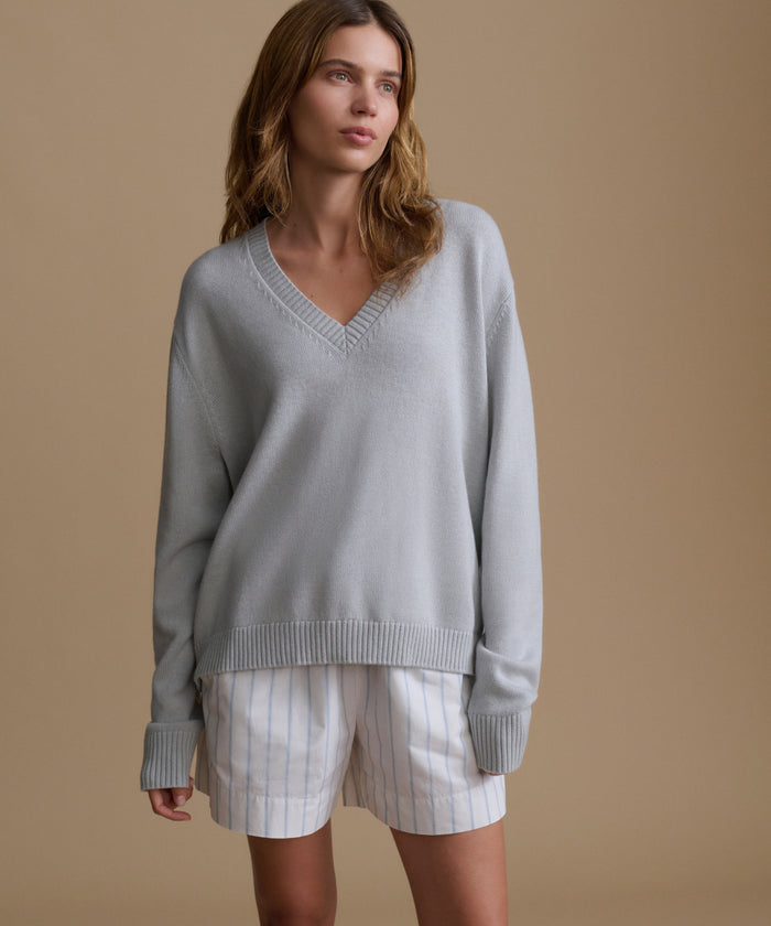 Woman wearing a light blue v-neck sweater with blue and white stripe shorts against a brown wall.