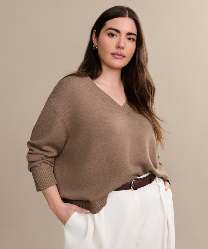 A woman with long brown hair exudes effortless style in the Simone V-Neck, a loose cotton-merino sweater, paired with white trousers and a brown belt, standing confidently against a beige background.