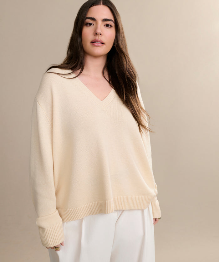 A woman with long brown hair wears the Simone V-Neck in ivory fine cotton merino with rolled-up sleeves and white pants, standing against a plain beige background.