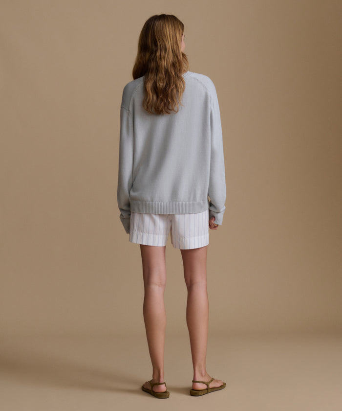 Woman wearing a light blue sweater with blue and white stripe shorts against a brown wall.