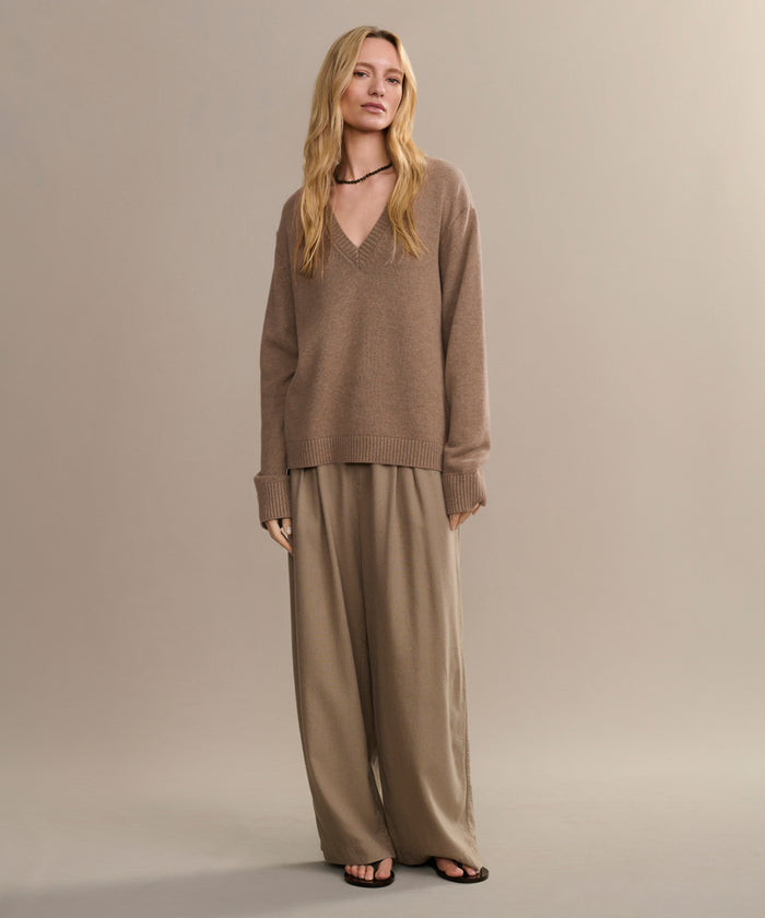 A person with long, wavy blonde hair and a neutral expression stands against a plain background wearing the Simone V-Neck sweater, wide-legged beige trousers, black sandals, and a black choker necklace.