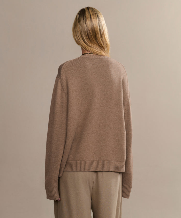 A person with blonde hair, wearing the loose-fitting Simone V-Neck brown sweater and beige pants, stands with their back to the camera against a plain beige background.