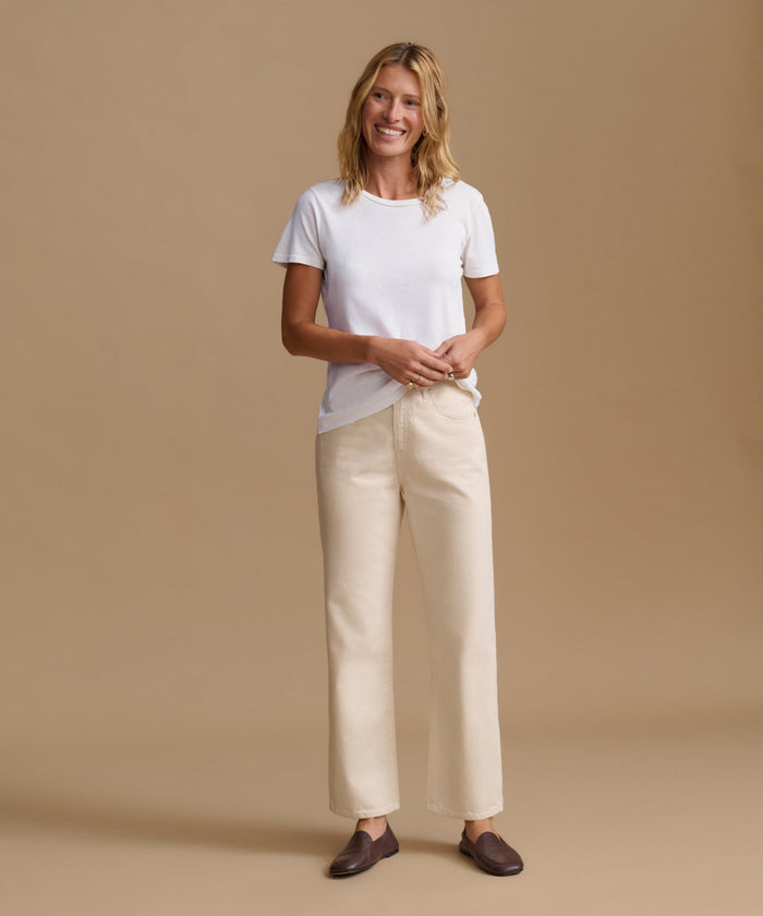 A woman modeling a white short-sleeve crewneck t-shirt and cream wide-leg trousers, paired with dark brown loafers, against a solid beige studio background.